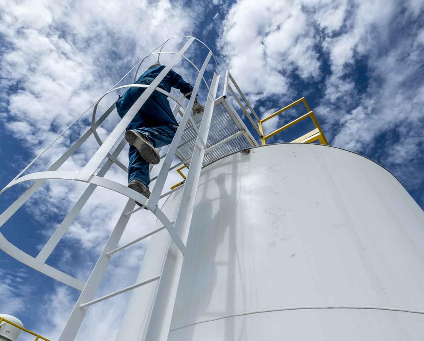 一名男子爬上一个储油罐上的梯子，从下面拍摄，抬头望向云层密布的天空