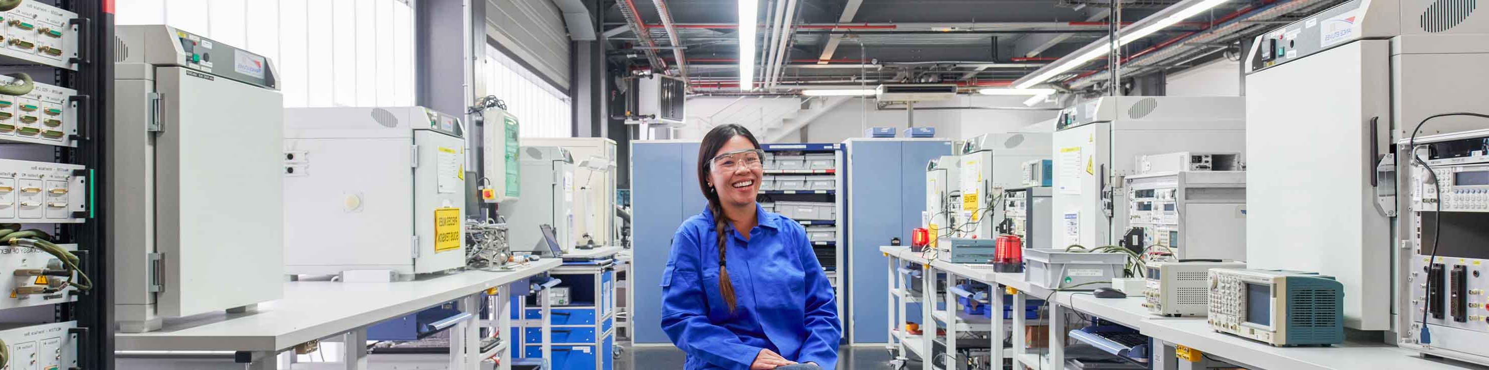 戴着护目镜，穿着黑裤子和蓝色实验服的女人坐在实验室里仍然微笑着
